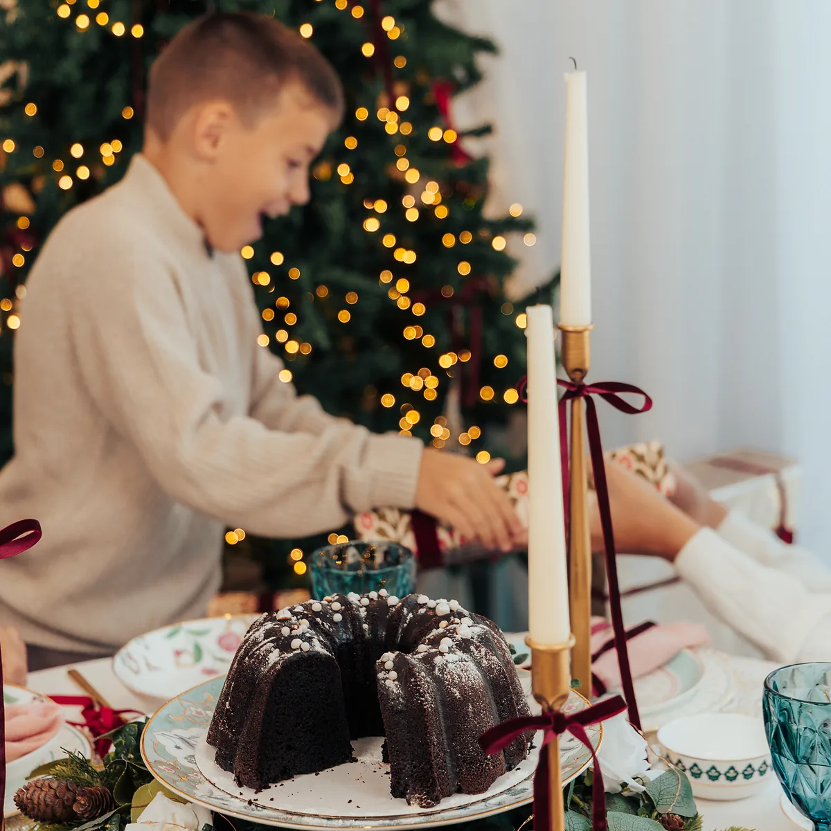 A dusted chocolate bundt cake on the table, a boy reaching across behind it, tree lights out of focus