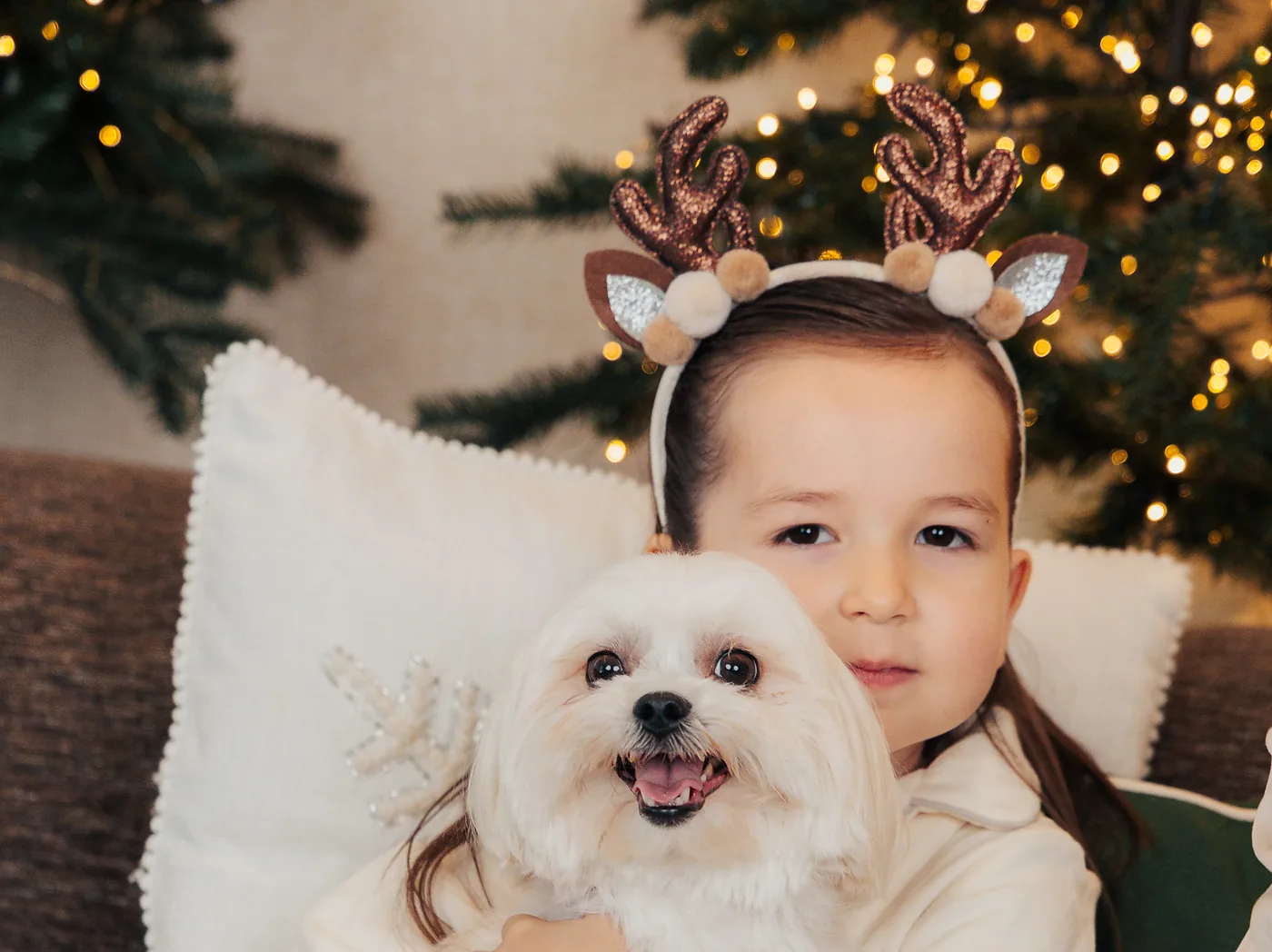 A girl in reindeer antlers holding her small white dog on the sofa in front of the tree