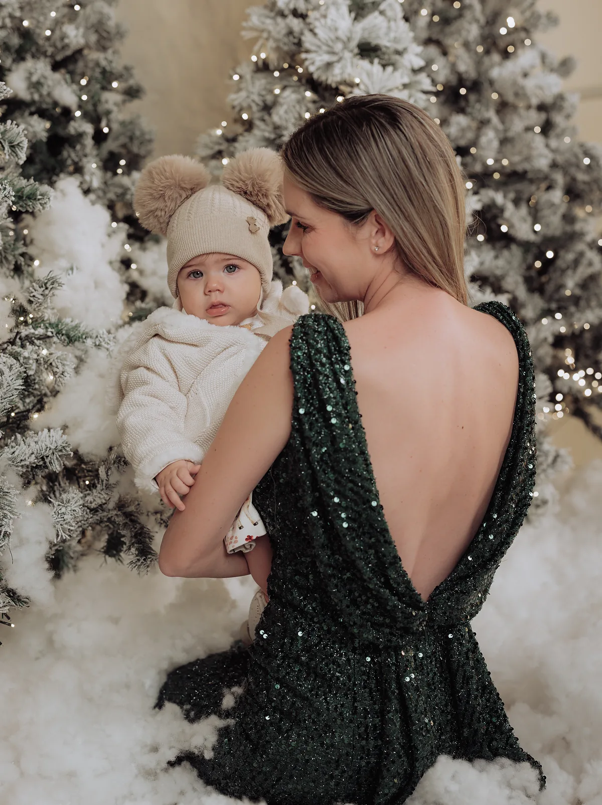 A mother in a deep green sequin dress turning to look at the baby she is holding, between two snowy trees