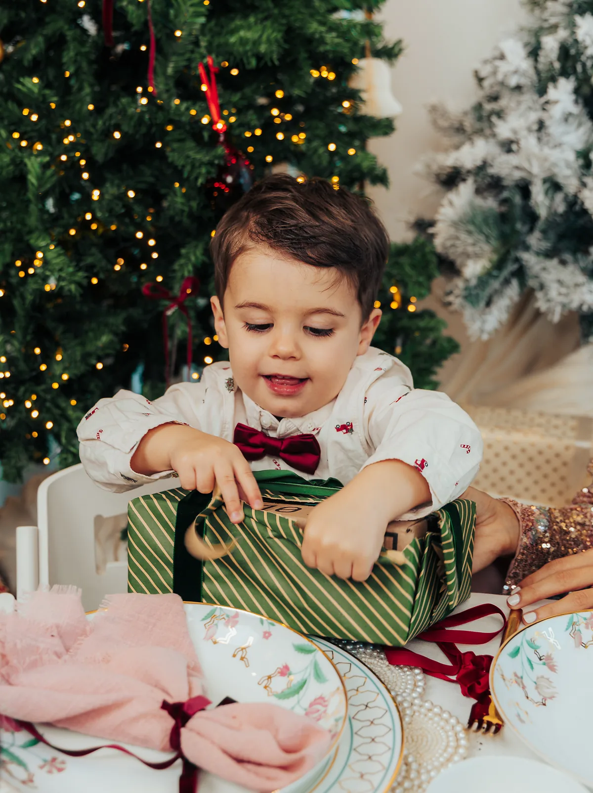 A small boy in a burgundy bow tie pulling the ribbon off a green striped present at the table