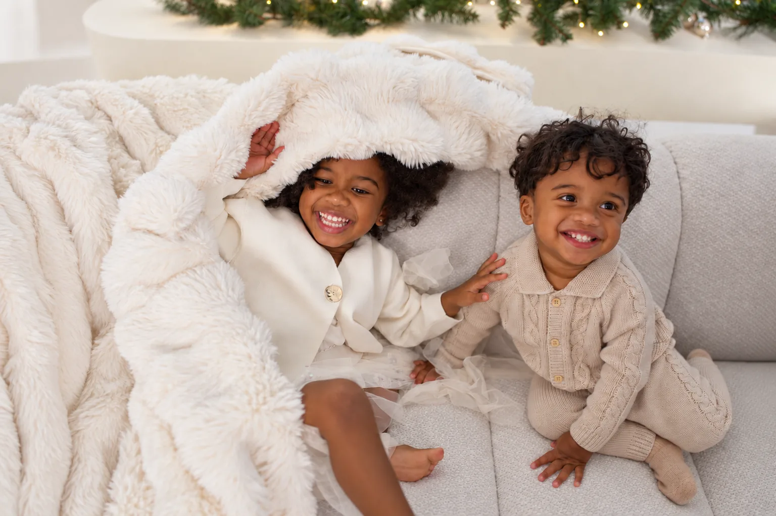 Two small children laughing on a sofa under a soft cream blanket, a lit garland behind them