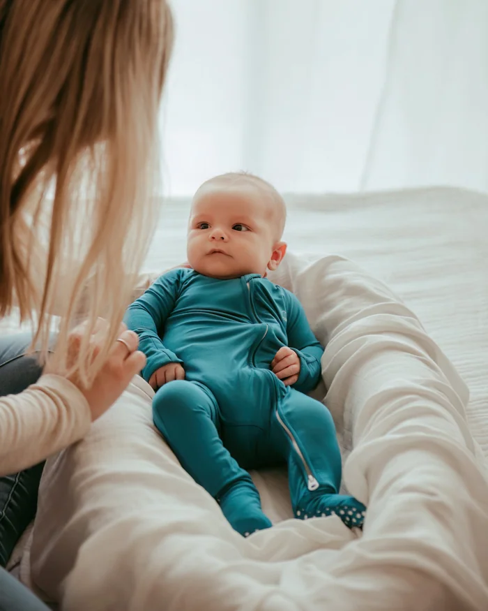A baby in a teal sleepsuit lying in a soft nest while a parent watches