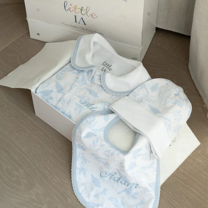 An open gift box of pale blue printed baby clothes and a bib embroidered with a name