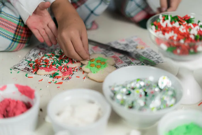 A child's hands pressing sprinkles onto an iced biscuit beside bowls of red and green sugar