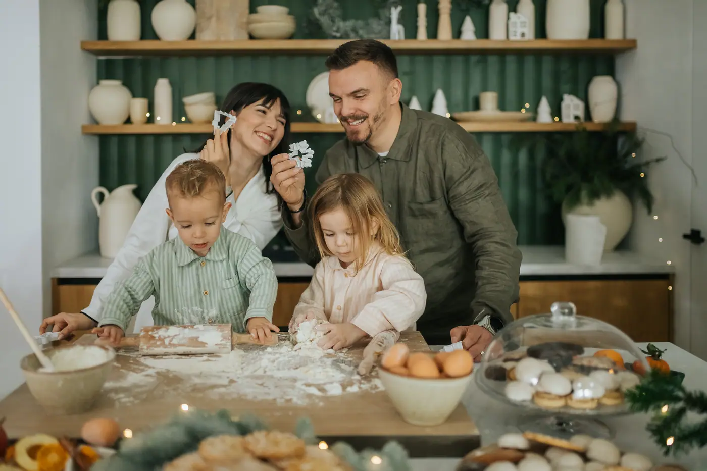 A mother and father laughing as their son and daughter press dough on a floured counter, a green kitchen shelf of white jugs behind them