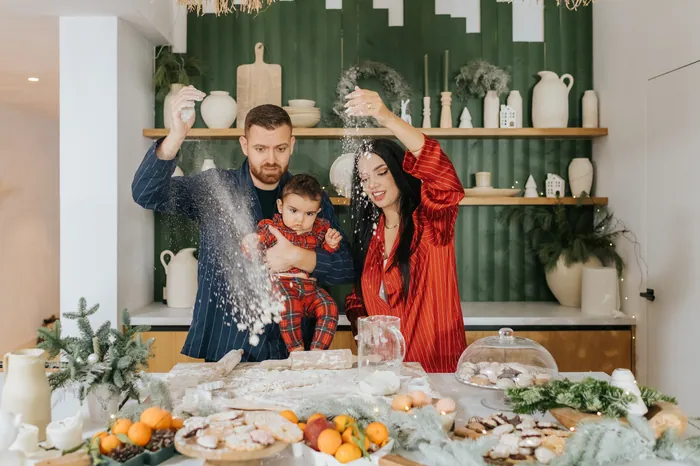 A father holding a baby in red tartan pyjamas while he and the mother throw flour into the air over a kitchen counter laid with biscuits and clementines