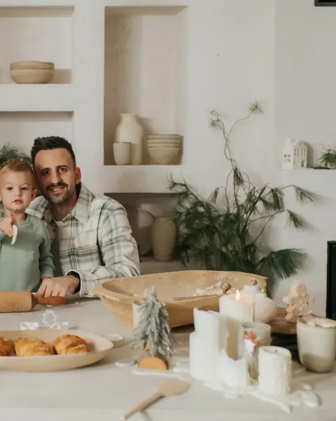 A family of three smiling behind a laid table with pillar candles, a cake dome, croissants and a bowl of fruit, greenery on the shelves behind them