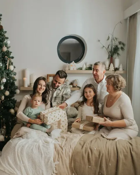 Grandparents, parents and three children opening presents together on a bed beside a tall decorated Christmas tree