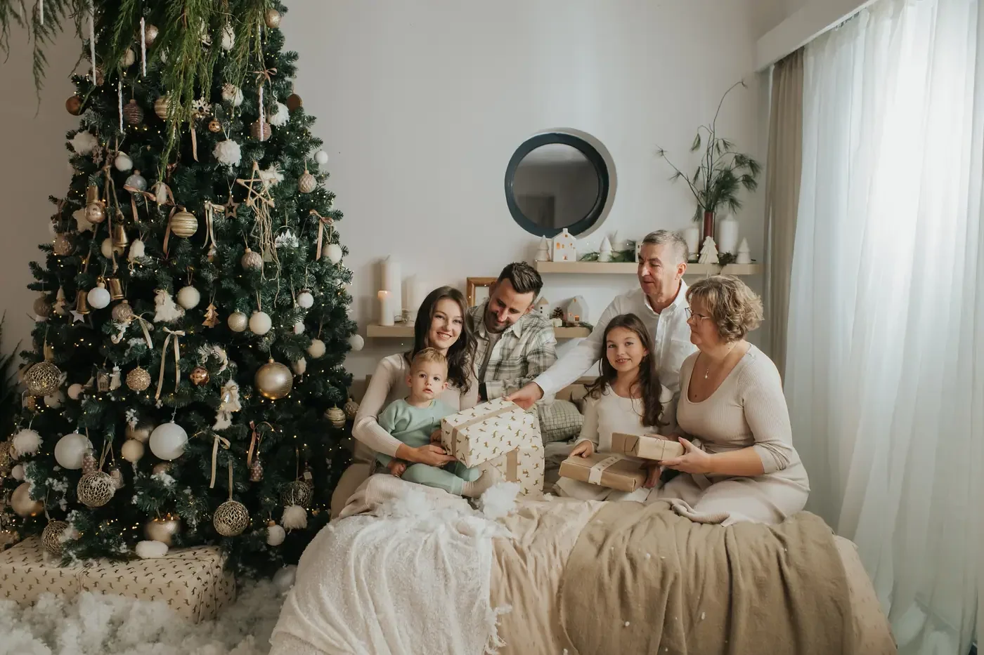 Grandparents, parents and three children opening presents together on a bed beside a tall decorated Christmas tree