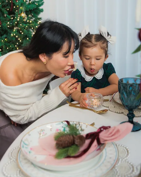 A mother and her small daughter in a tartan dress leaning over a candle at a laid Christmas table