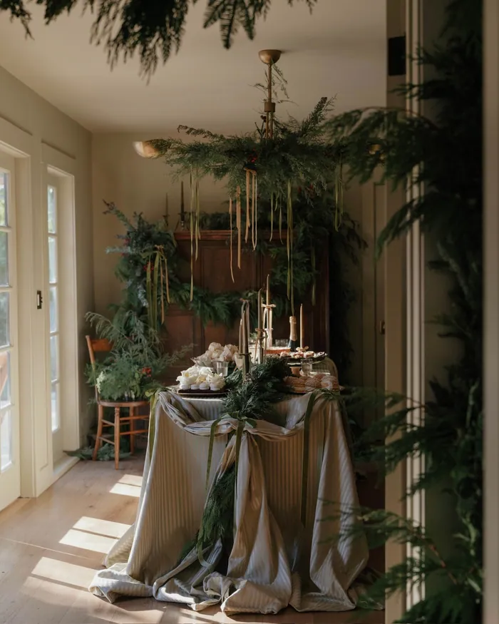 A table draped in grey linen and set for a festive meal, greenery hanging above it in a sunlit room