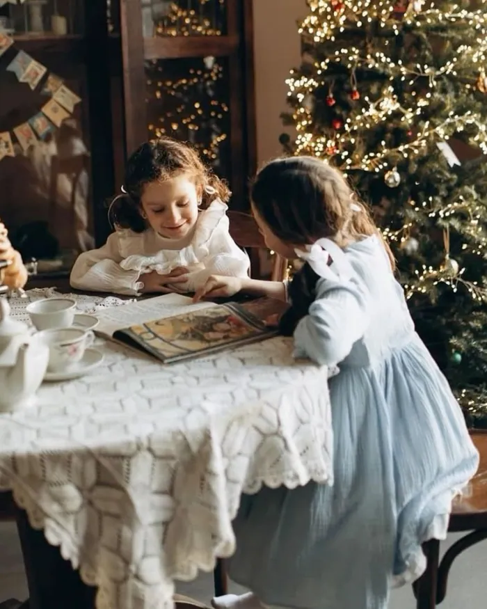 Two little girls at a round table with a lace cloth, looking at a book beside a lit Christmas tree