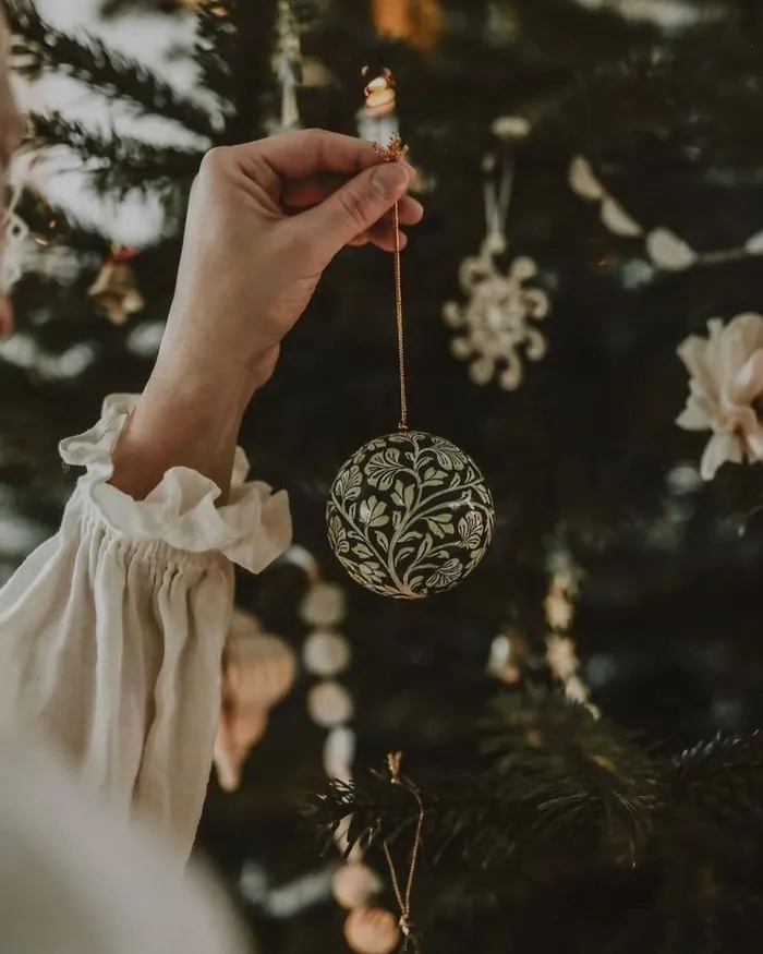 A hand in a ruffled sleeve hanging a round ornament on a Christmas tree in the dark