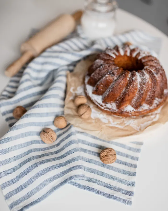 A bundt cake dusted with sugar on a striped linen cloth, walnuts and a rolling pin beside it