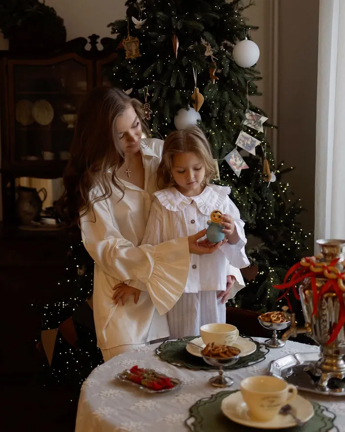 A mother and her small daughter in cream pyjamas at a laid table beside the Christmas tree