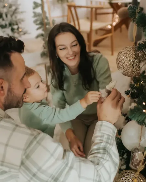 A family decorating a flocked Christmas tree together, the little boy reaching up to hang a bauble
