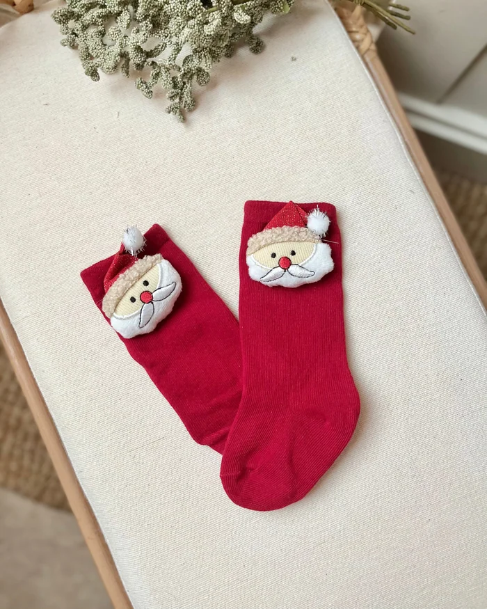 A pair of red baby socks with Father Christmas faces on the cuffs, on a cream cushion