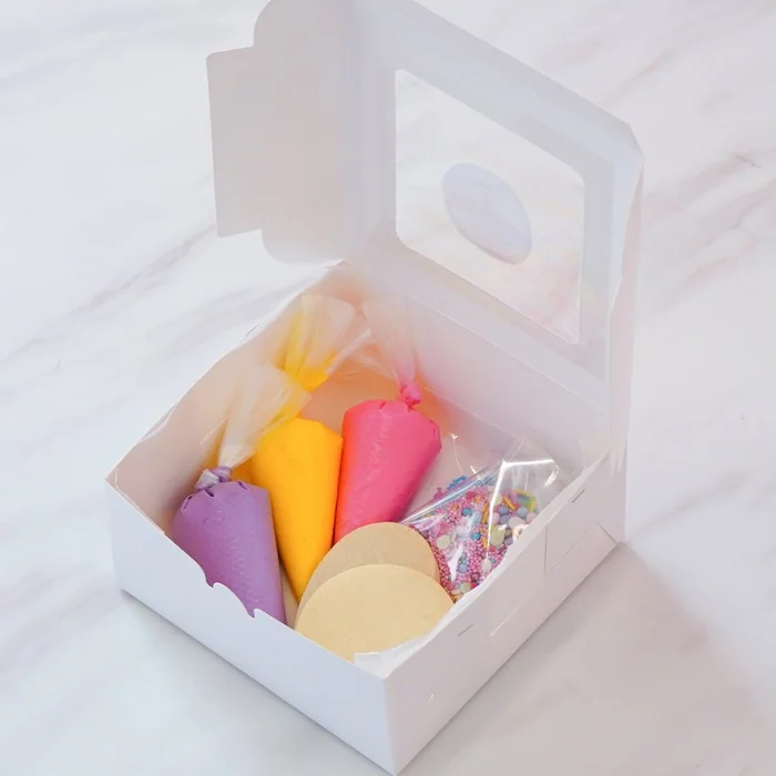 A cookie decorating kit in an open white box: piping bags of icing, sprinkles and plain round biscuits