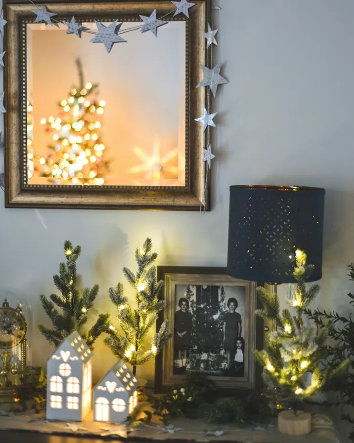 A framed black-and-white family photograph among small lit Christmas trees and paper houses, a mirror hung with a star garland above