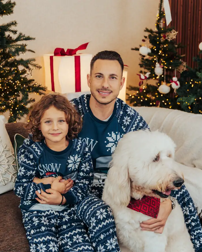A father and his son in navy fair isle pyjamas on the sofa with their big white doodle in a red bandana