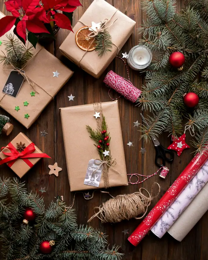 Presents wrapped in brown paper and twine with sprigs of pine, dried orange and red baubles, beside rolls of ribbon and a poinsettia
