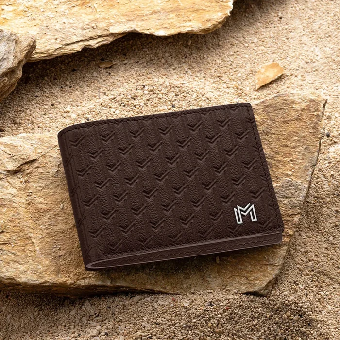 A dark brown embossed leather wallet with a small metal M, resting on sandstone