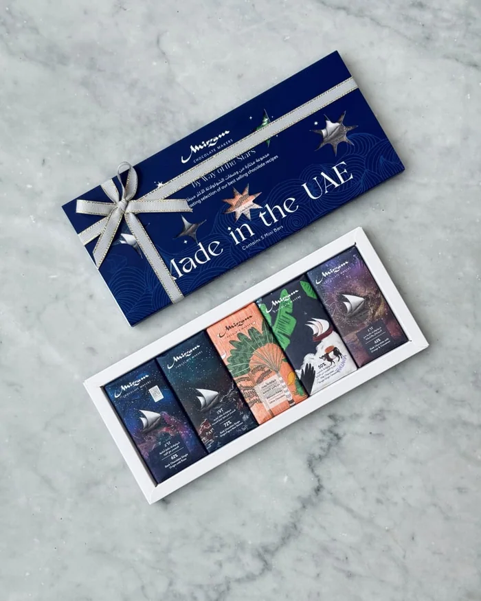 A navy Mirzam gift box tied with a ribbon above an open box of chocolate bars, on marble