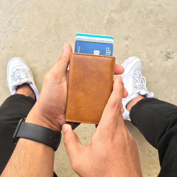 Hands holding a tan leather card holder with blue cards sliding out of the top