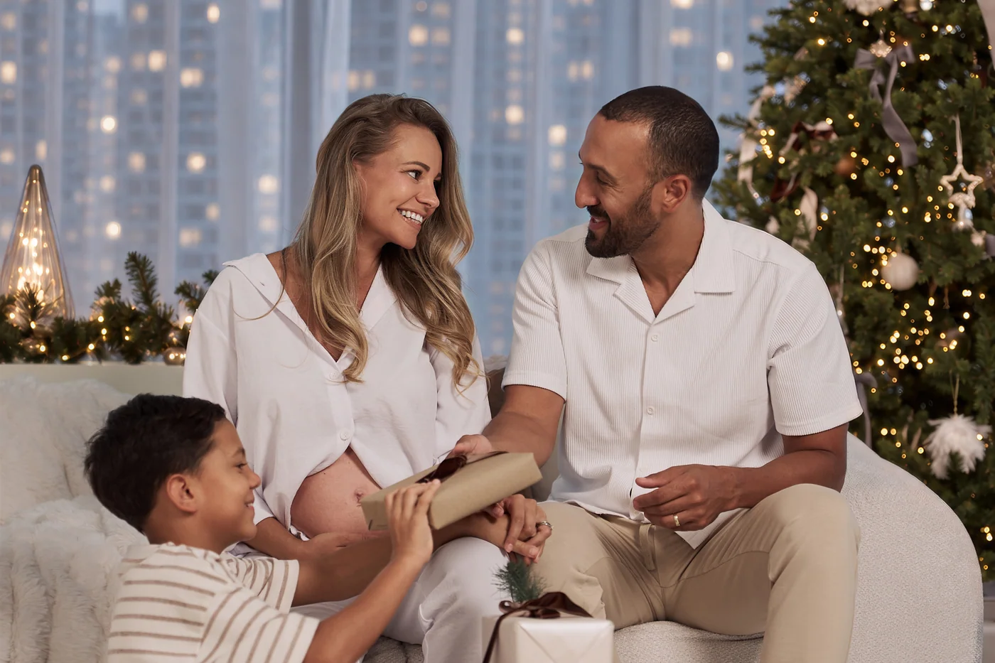 A smiling father in a checked shirt with his two sons in front of a lit Christmas tree and city windows