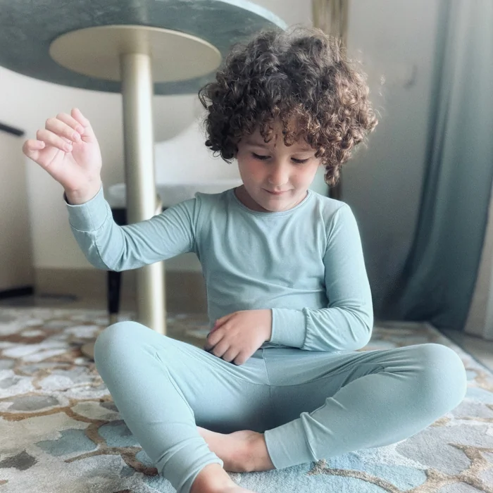 A curly-haired boy in soft light blue pyjamas sitting cross-legged on a rug