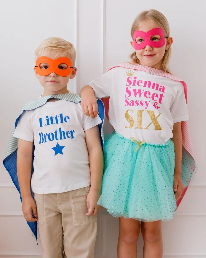 A brother and sister in superhero masks and capes, wearing printed name T-shirts