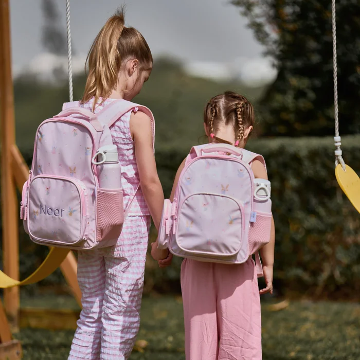 Two girls holding hands by a swing, both wearing pink fairy print backpacks, one embroidered with the name Noor