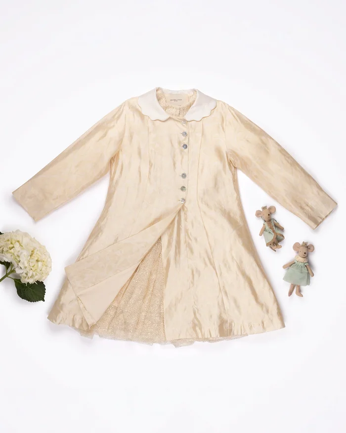 A girl's cream silk coat dress with a round collar, laid flat beside white flowers