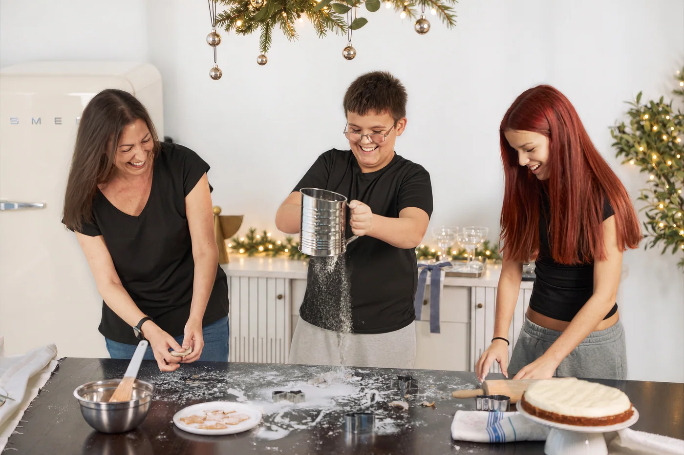 A mother and two teenagers baking in a white Christmas kitchen, the boy sifting flour and the girl rolling dough
