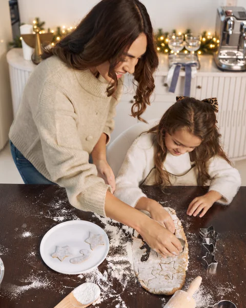 A mother and daughter rolling out biscuit dough together at a floury kitchen table, fairy lights behind them