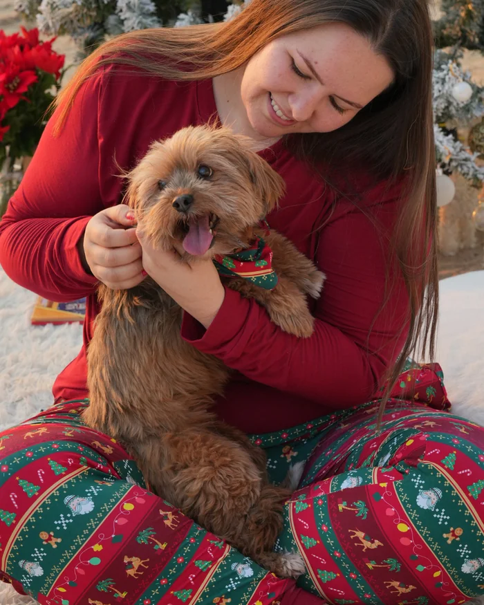 A woman in a red top and Christmas print pyjama trousers cuddling a small fluffy dog