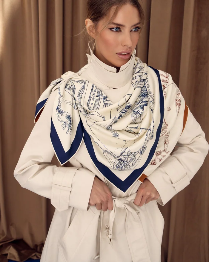 A woman in a cream trench coat wearing a silk scarf printed with Abu Dhabi heritage scenes in navy on ivory