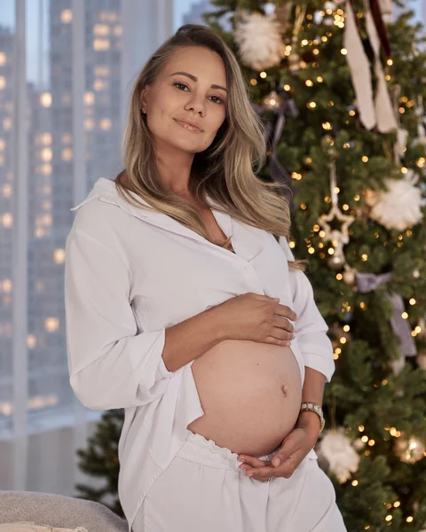A pregnant woman in white smiling beside a decorated Christmas tree, city windows behind