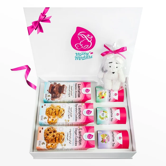 An open white Milky Makers gift box with a pink ribbon, holding boxes of lactation cookies, brownies and tea and a small knitted toy