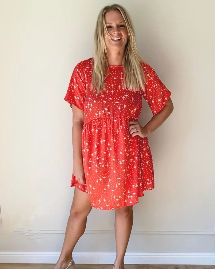 A smiling woman in a short red dress printed with white snowflakes, standing against a pale wall