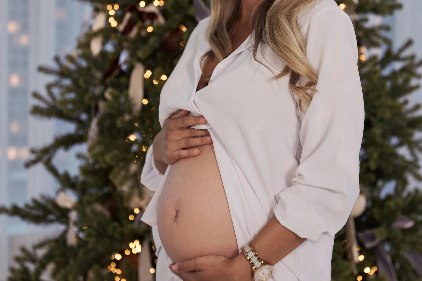 A pregnant mum in a white shirt holding her bump in front of a lit Christmas tree