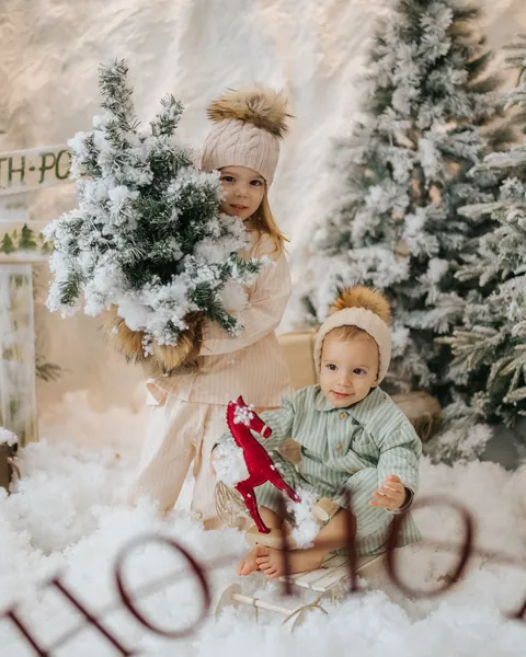Two small children in bobble hats in the snow among flocked trees, the older one carrying a little frosted tree
