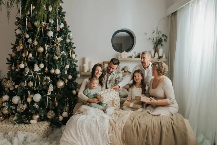 Grandparents, parents and three children opening presents together on a bed beside a tall decorated Christmas tree
