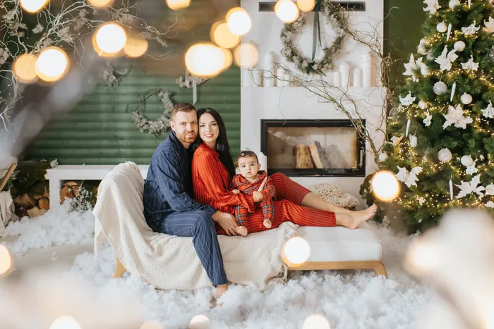 A mother and father in pyjamas on a snowy sofa beside a fireplace, their baby in red check on the mother's lap, Christmas lights glowing in the foreground