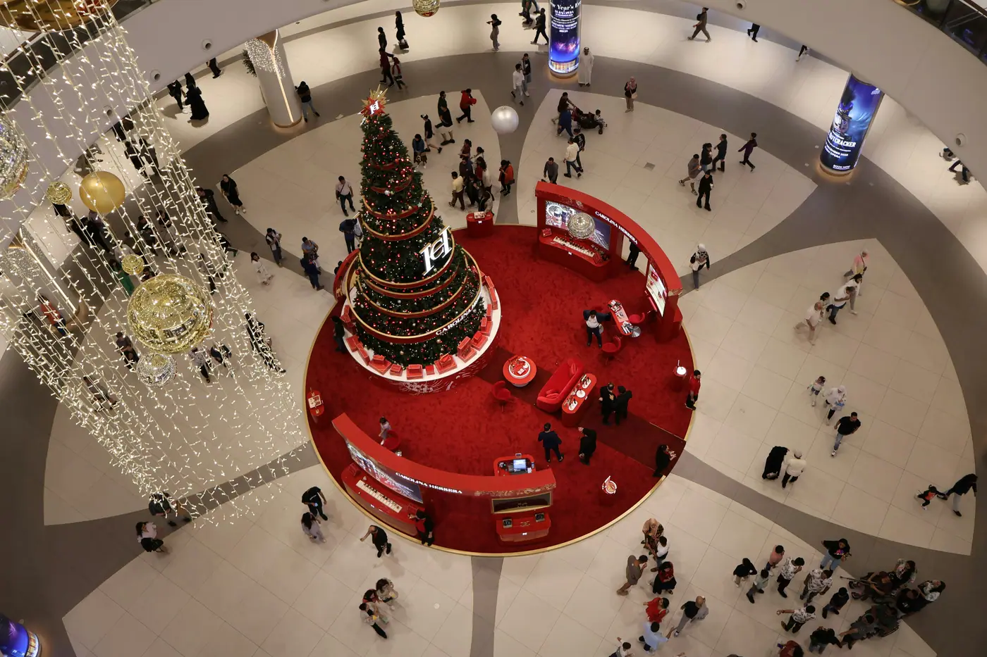 A giant Christmas tree on a red base in the middle of a Dubai mall atrium, shoppers circling it, seen from above