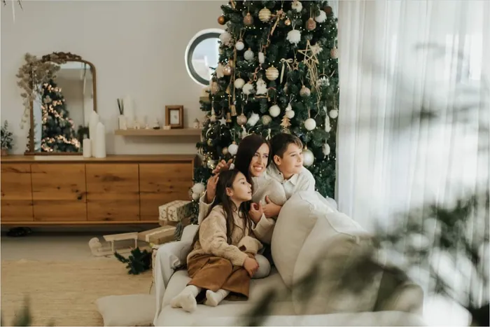 A mother and her two children curled up together on a white sofa beside a tall decorated tree, looking out of the window