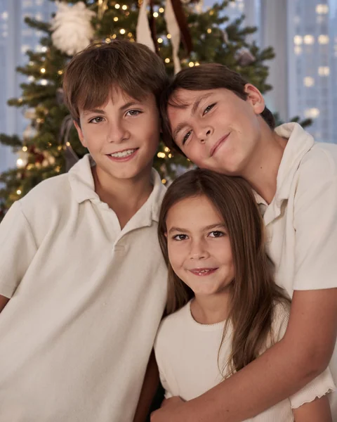 Two brothers and their little sister leaning together and smiling in front of a Christmas tree