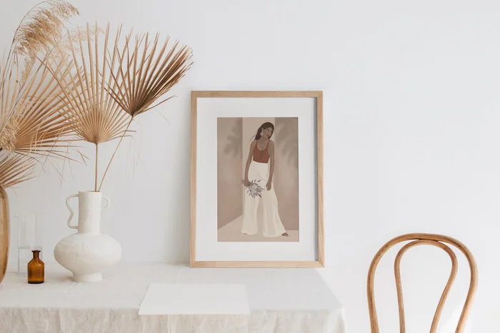 A framed illustration on a white table beside a vase of dried palm leaves and a bentwood chair