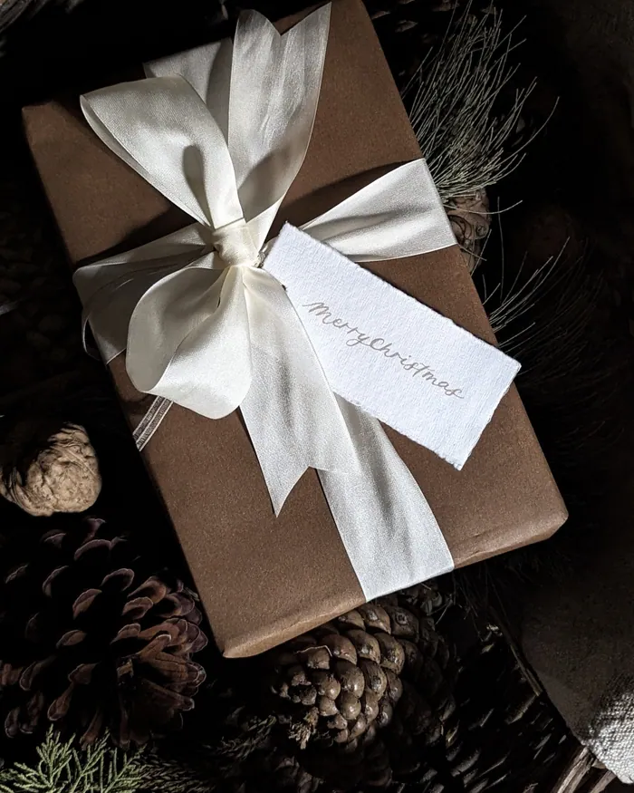A wrapped present tied with cream ribbon and a handwritten tag among pine cones