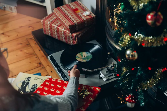 A hand lowering a record onto a turntable beside wrapped presents and a Christmas tree